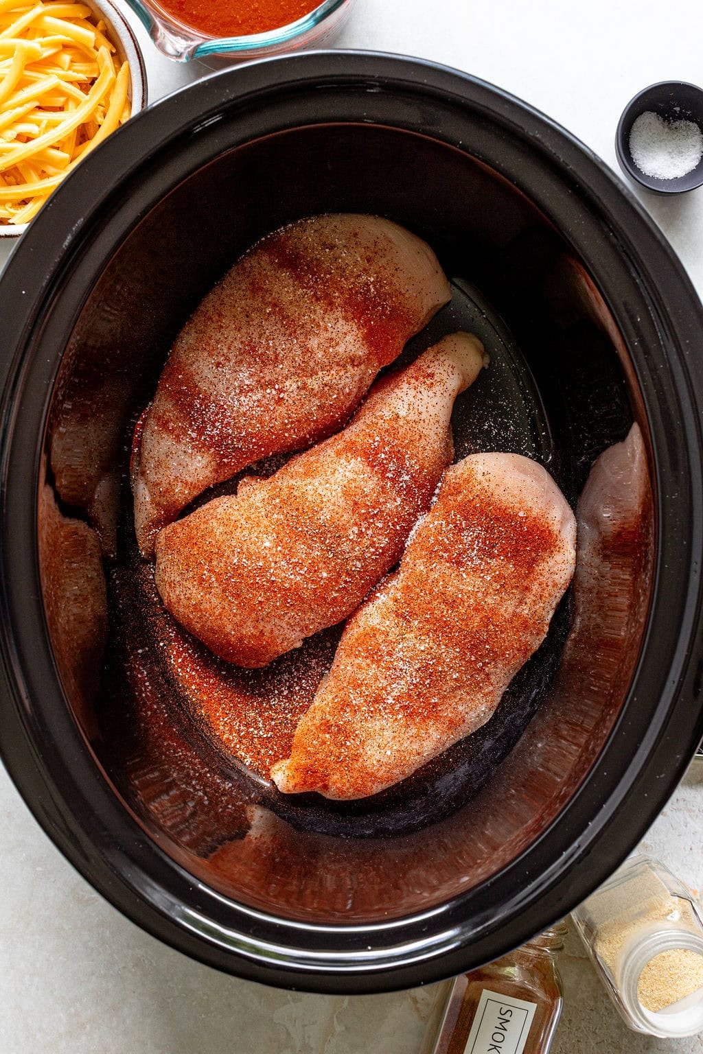 Three raw, seasoned chicken breasts in a slow cooker with shredded cheese, spices, and salt in bowls nearby on a countertop.