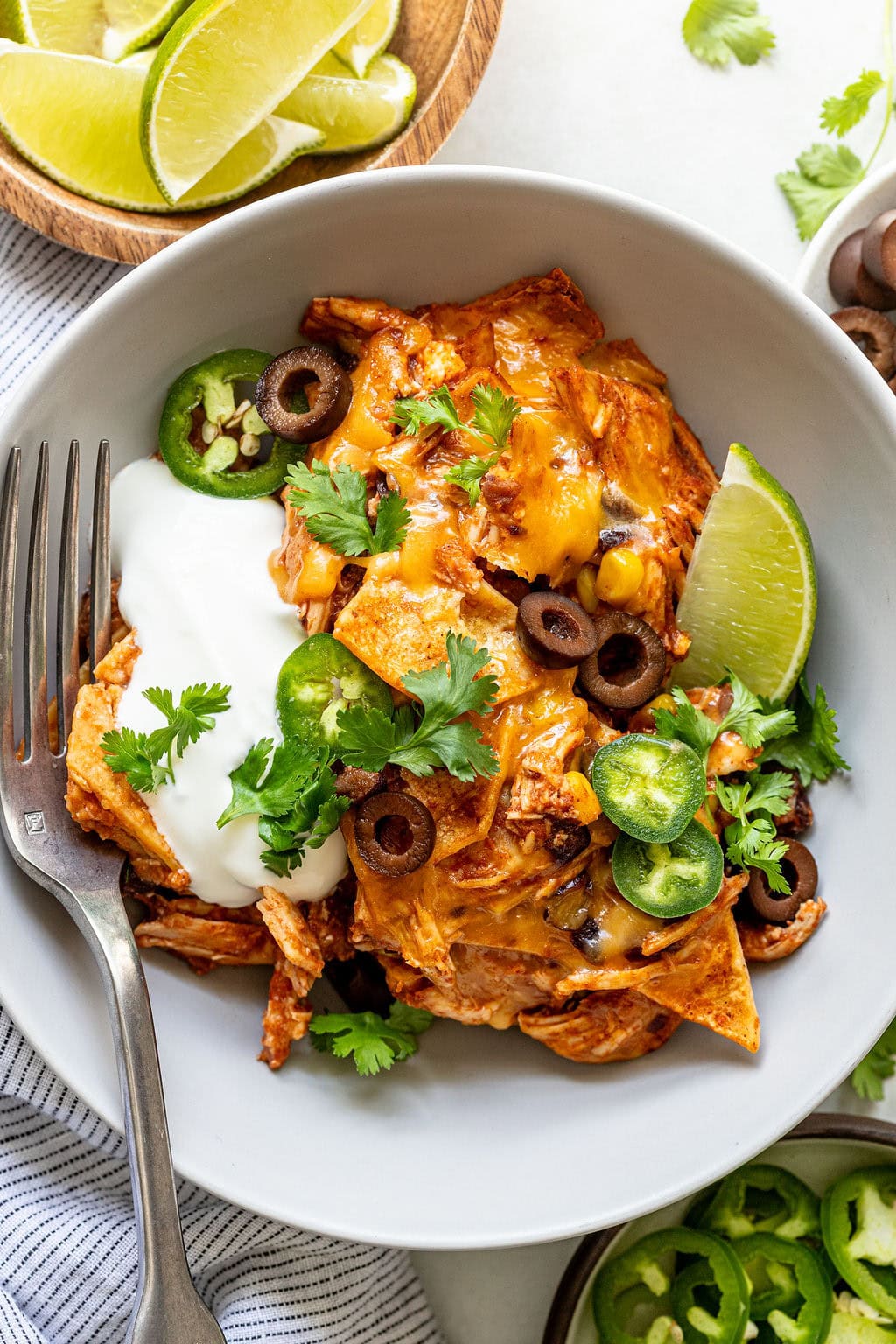 A bowl of chicken enchiladas topped with sour cream, jalapeños, sliced olives, cilantro, and a lime wedge, with lime slices and jalapeños on the side.