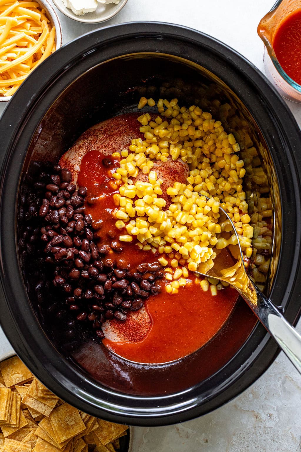 A slow cooker filled with black beans, corn, red sauce, and a spoon, surrounded by shredded cheese, sour cream, and chips.