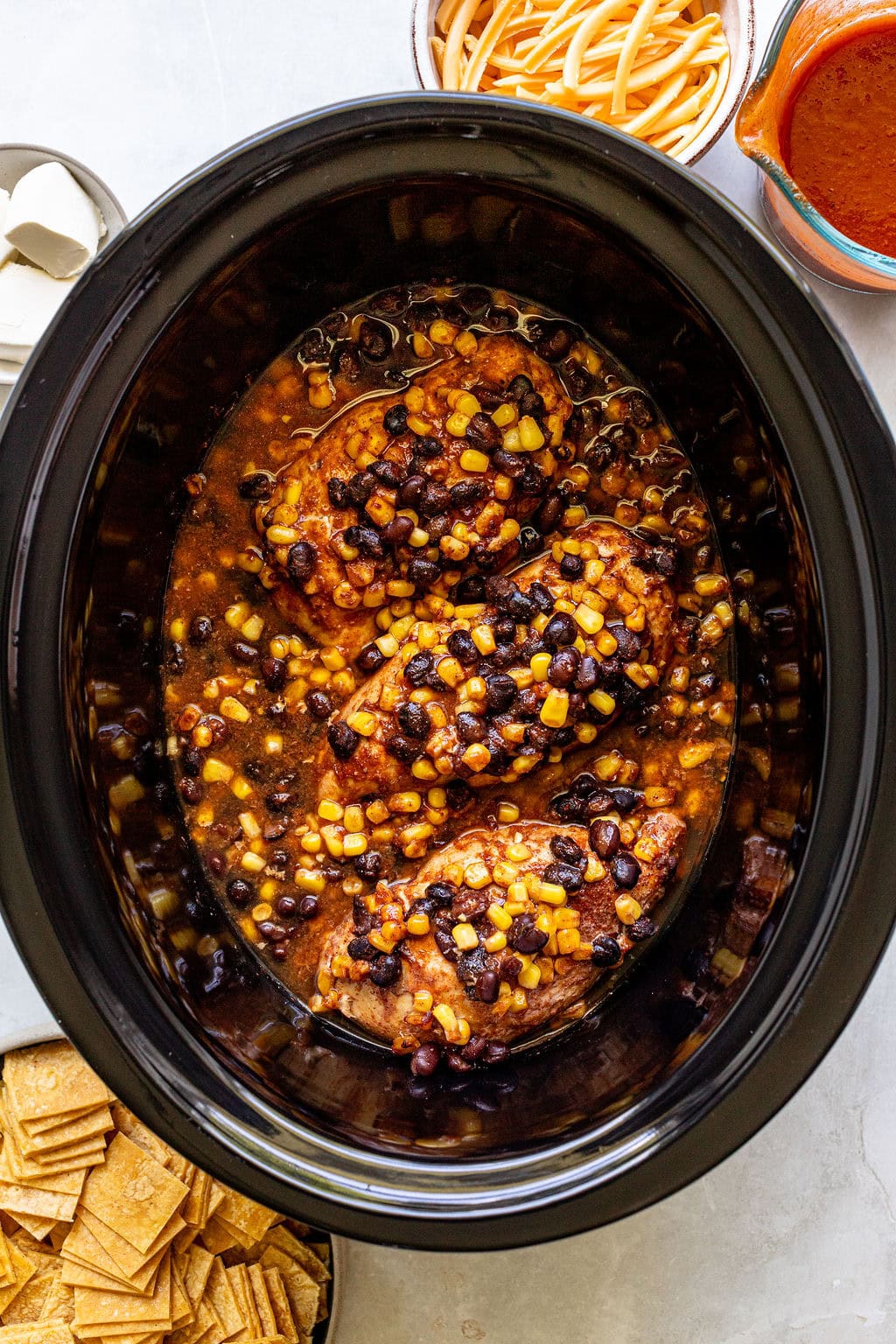 Three chicken breasts in a slow cooker with a sauce containing black beans and corn, surrounded by plates of cheese, sauce, and chips.