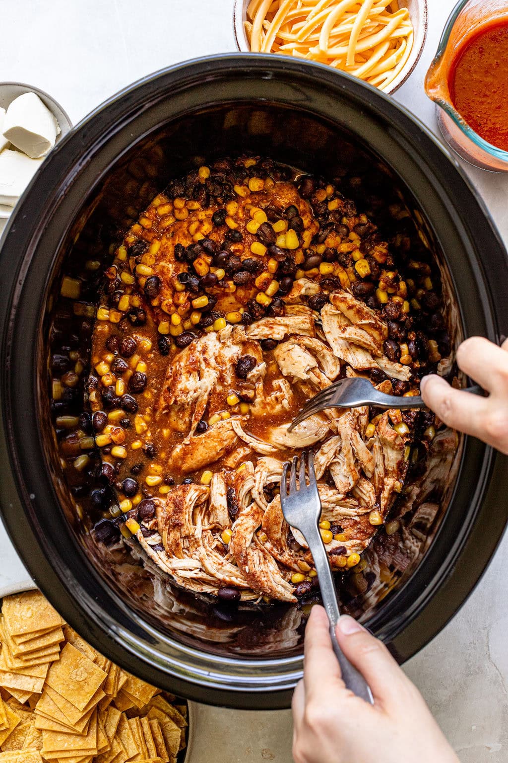 Two hands use forks to shred cooked chicken in a slow cooker with black beans, corn, and sauce.