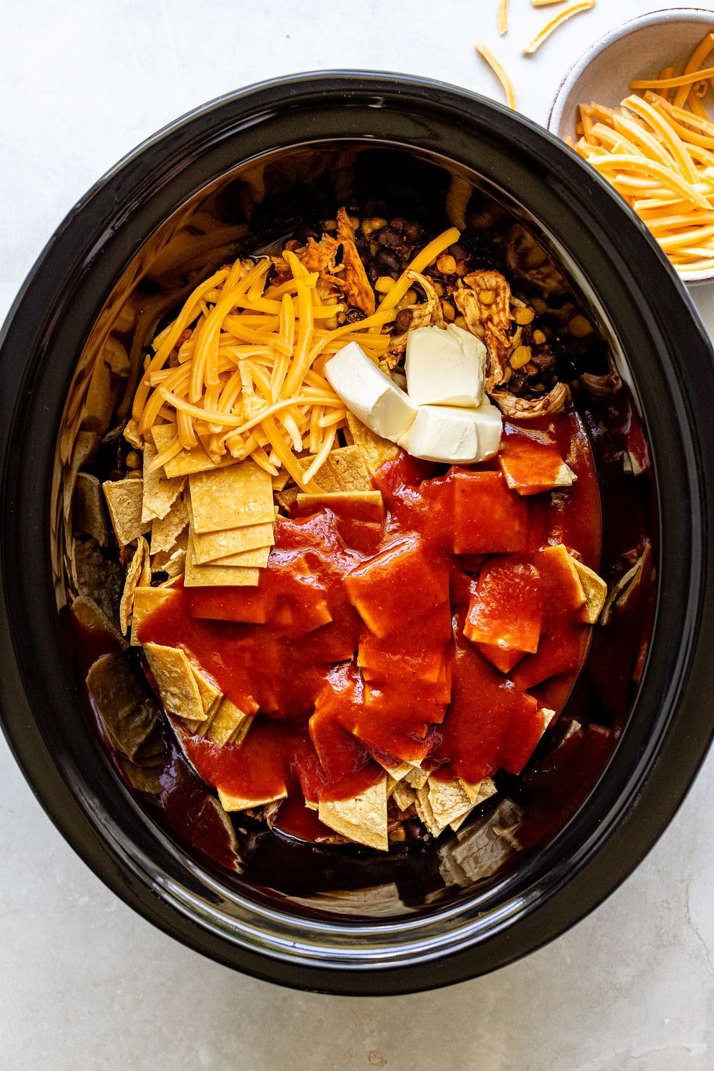 Ingredients for a casserole, including tortilla chips, shredded cheese, cream cheese, beans, and red sauce, are arranged inside a slow cooker before mixing or cooking.
