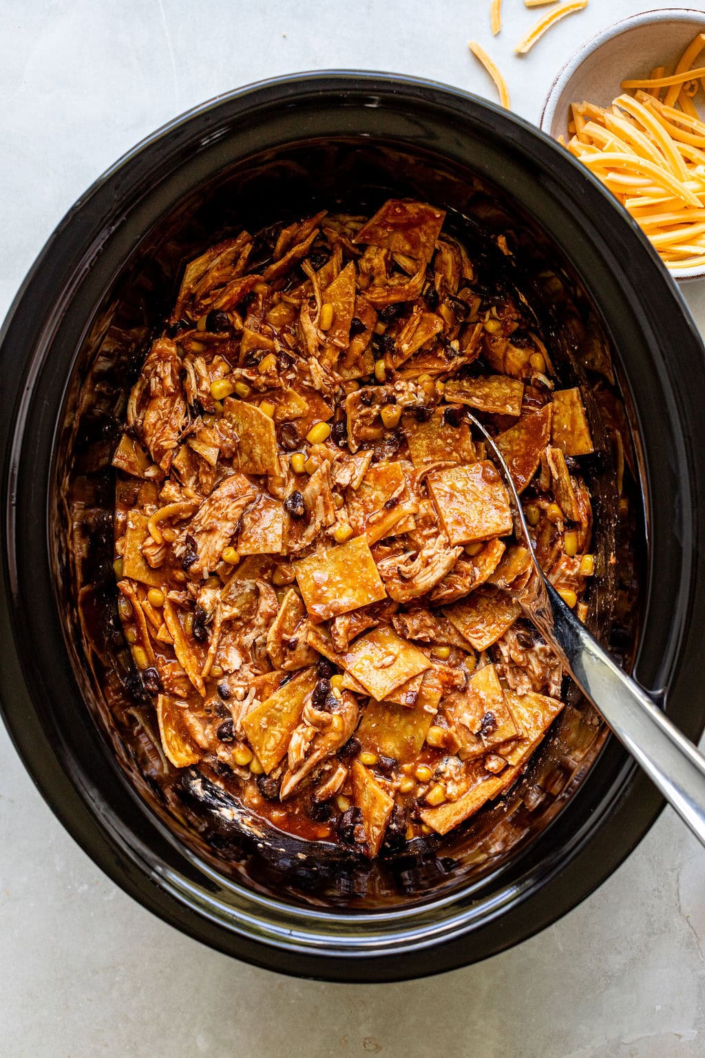 A slow cooker filled with shredded chicken, tortilla strips, black beans, corn, and sauce, with a serving spoon and a bowl of shredded cheese nearby.