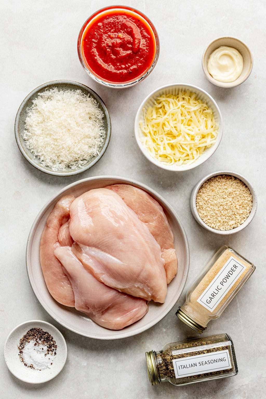 Raw chicken breasts on a plate surrounded by grated cheeses, marinara sauce, mayonnaise, breadcrumbs, garlic powder, Italian seasoning, salt, and pepper in small bowls on a white surface.