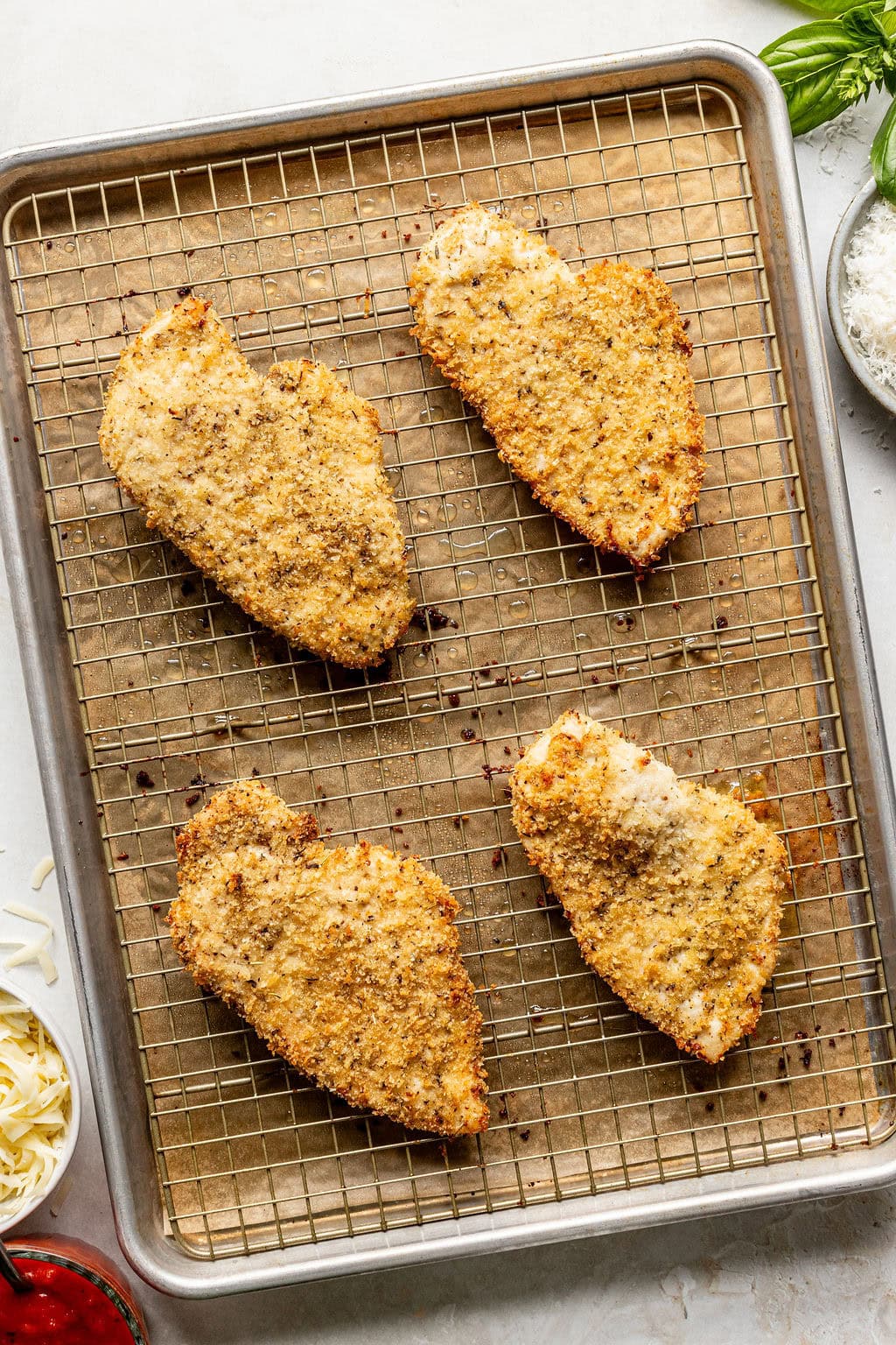 Four breaded chicken cutlets on a wire rack over a baking sheet, with bowls of shredded cheese and herbs nearby.