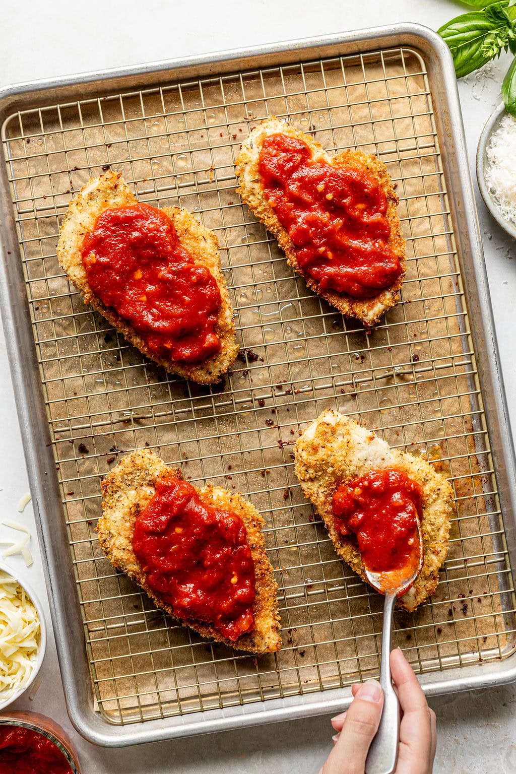 Four breaded chicken cutlets on a wire rack, three topped with red marinara sauce, and a hand spooning sauce onto the fourth cutlet.