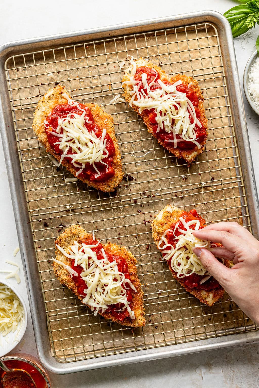 Four breaded chicken breasts on a wire rack with tomato sauce and shredded cheese, with a hand adding cheese to one piece.