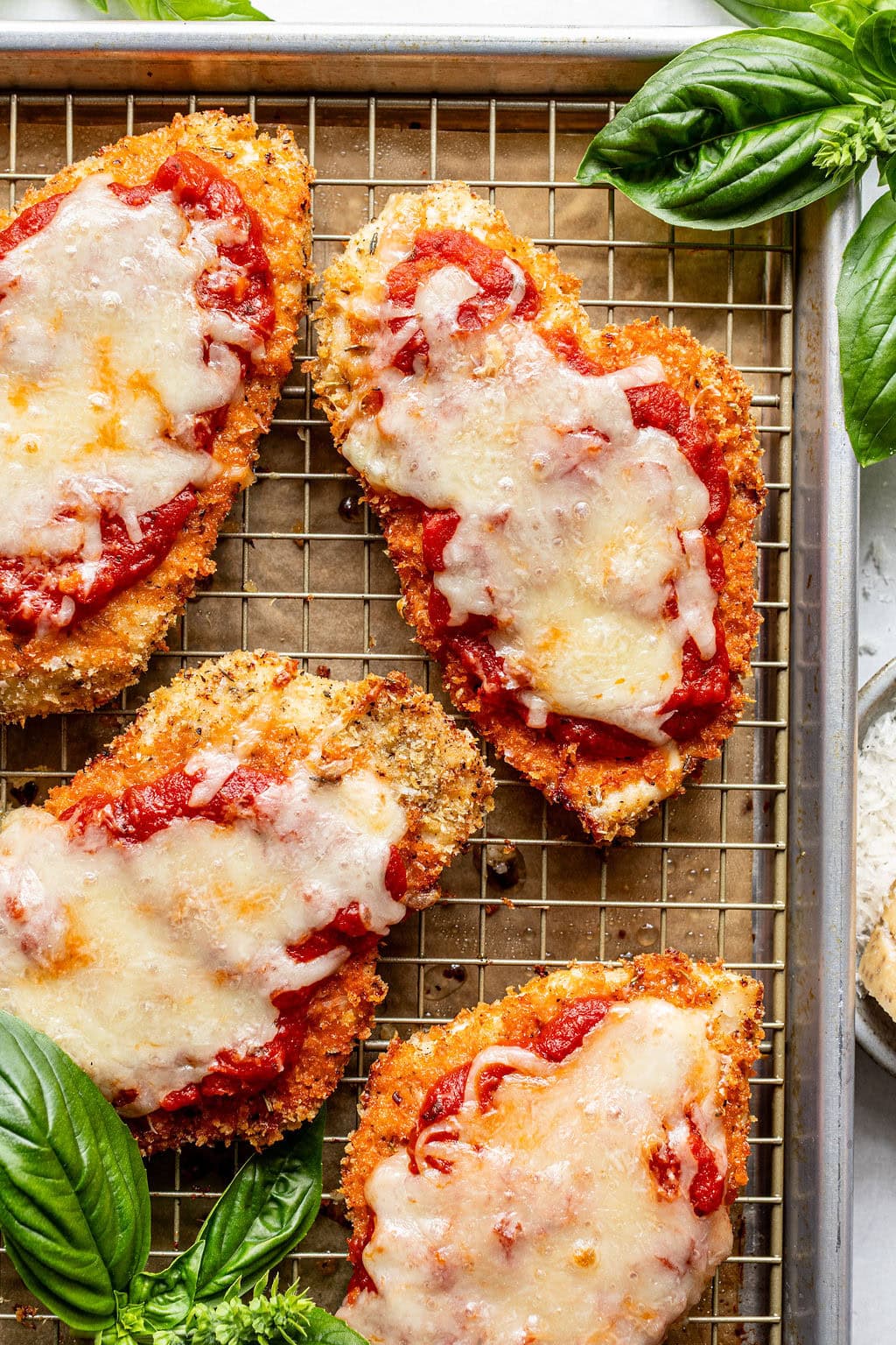 Four breaded chicken cutlets topped with tomato sauce and melted cheese on a wire rack, with fresh basil leaves nearby.