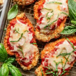 Breaded chicken cutlets topped with marinara sauce, melted cheese, and shredded basil, arranged on a wire rack with fresh basil leaves beside them.