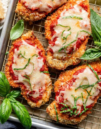Breaded chicken cutlets topped with marinara sauce, melted cheese, and shredded basil, arranged on a wire rack with fresh basil leaves beside them.