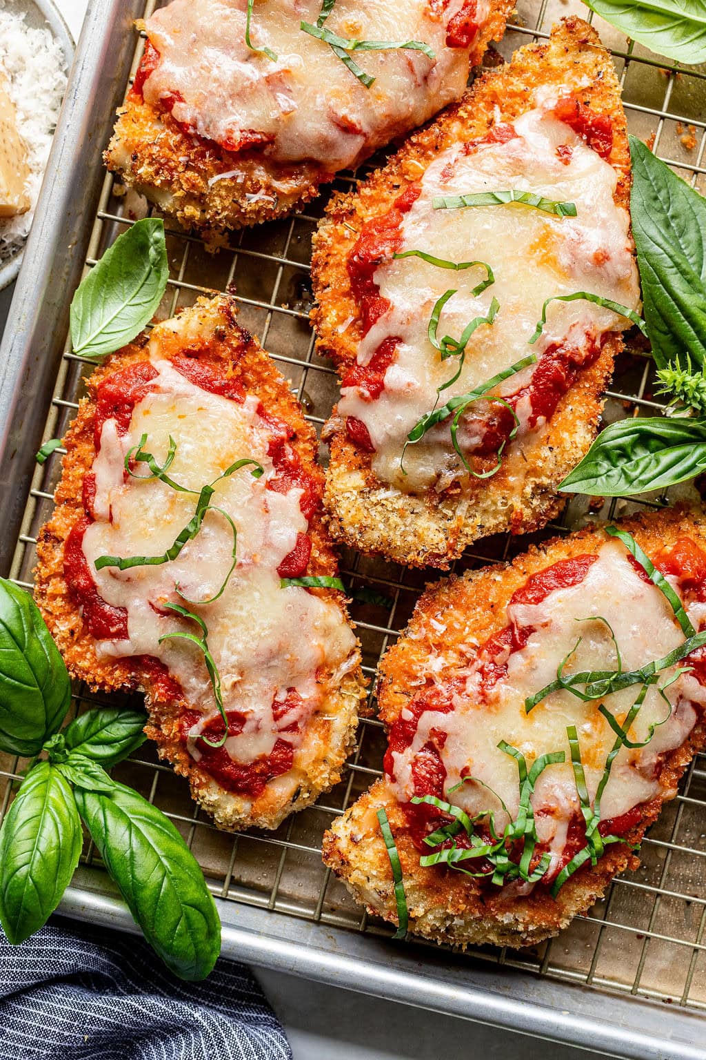 Breaded chicken cutlets topped with marinara sauce, melted cheese, and shredded basil, arranged on a wire rack with fresh basil leaves beside them.