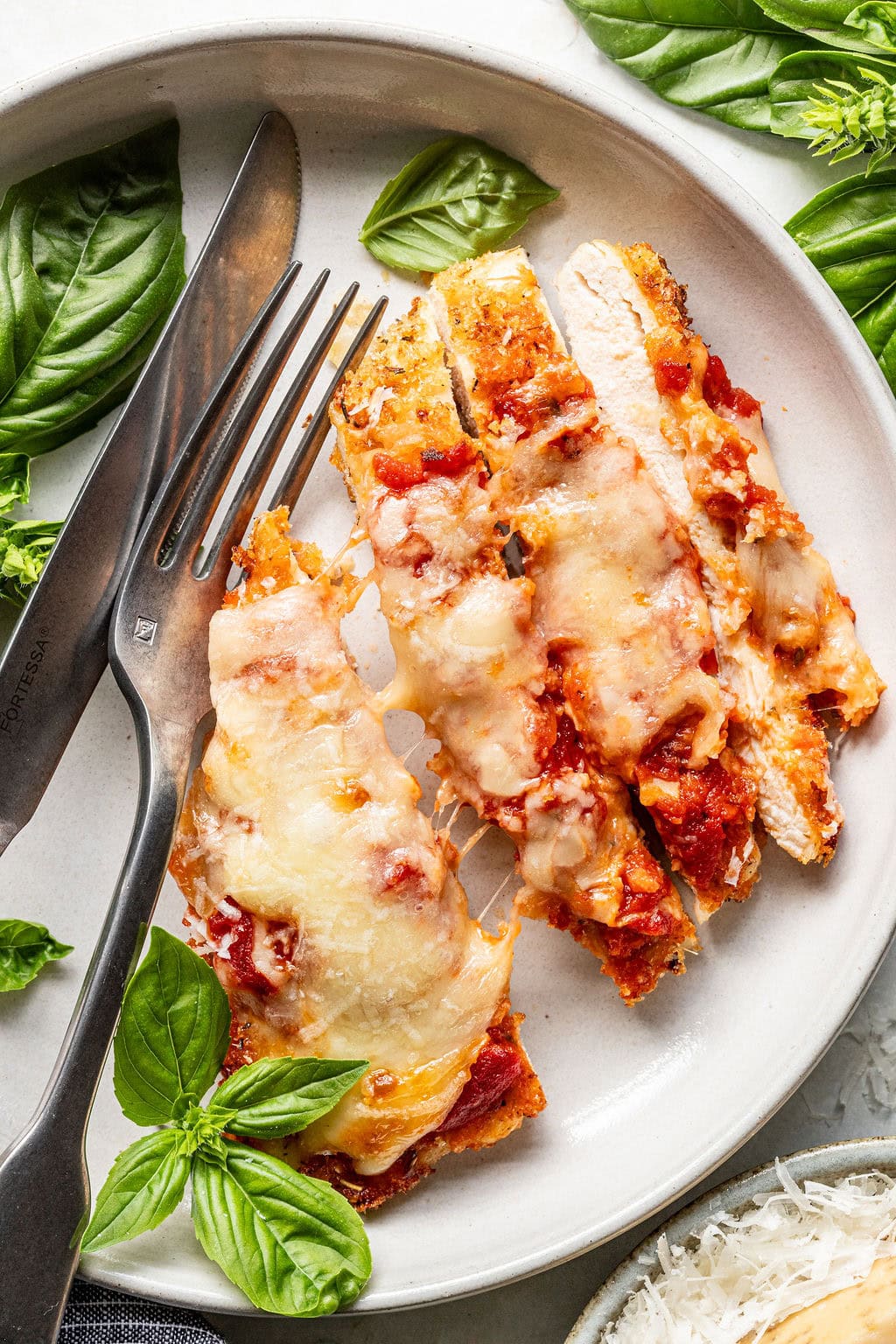 Sliced chicken parmesan topped with melted cheese and tomato sauce on a white plate, garnished with fresh basil leaves, with a fork and knife beside the chicken.