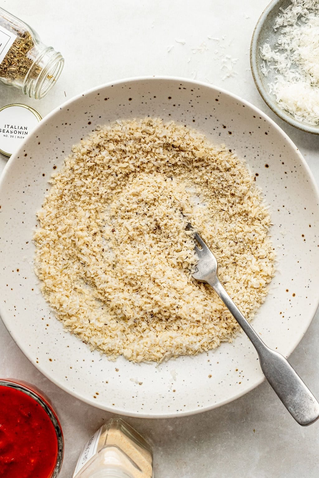 A speckled plate with seasoned breadcrumbs and a fork, surrounded by jars of spices, grated cheese, and a bowl of red sauce.