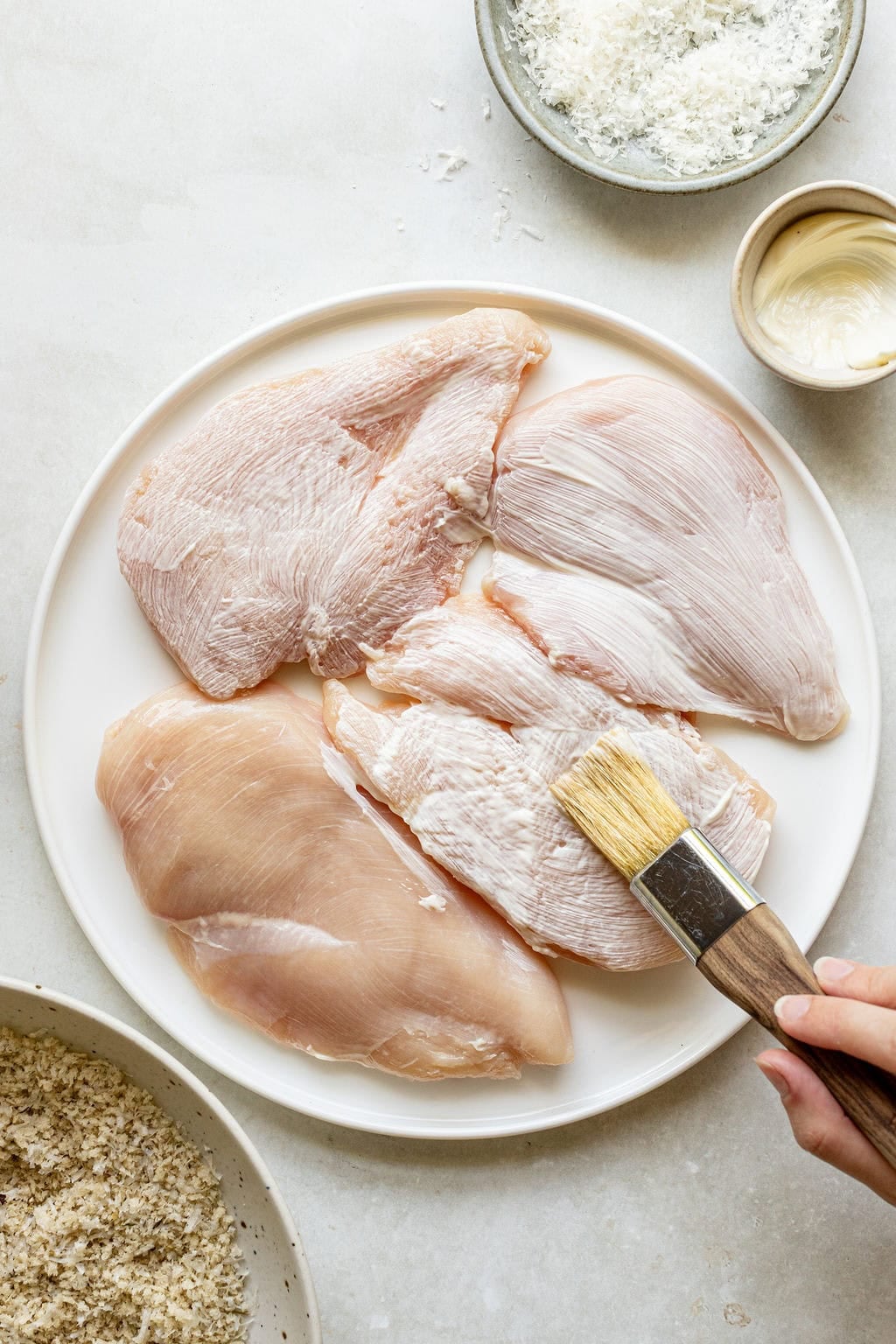A hand brushes raw chicken breasts on a white plate with sauce; bowls of bread crumbs and shredded cheese are nearby.