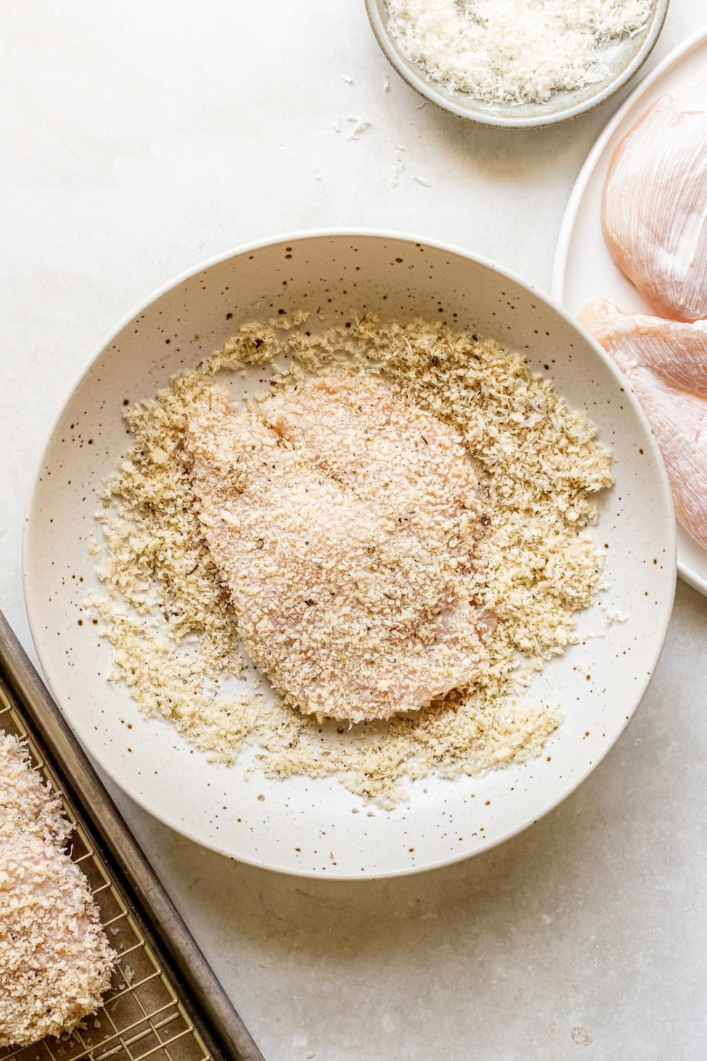 A raw chicken breast is being coated in breadcrumbs on a speckled plate, with more breadcrumbs, raw chicken, and a baking rack visible nearby.