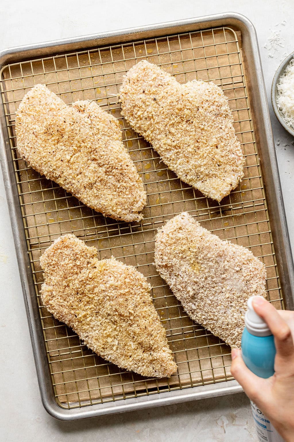 Four breaded chicken breasts on a wire rack over a baking sheet, with a hand spraying oil over them.