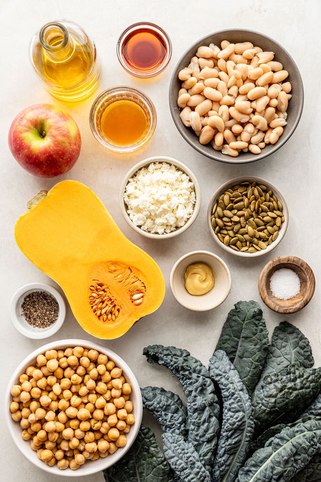 Assorted ingredients including kale, butternut squash, apple, white beans, chickpeas, feta, pepitas, oil, vinegar, mustard, salt, and pepper arranged on a light surface.