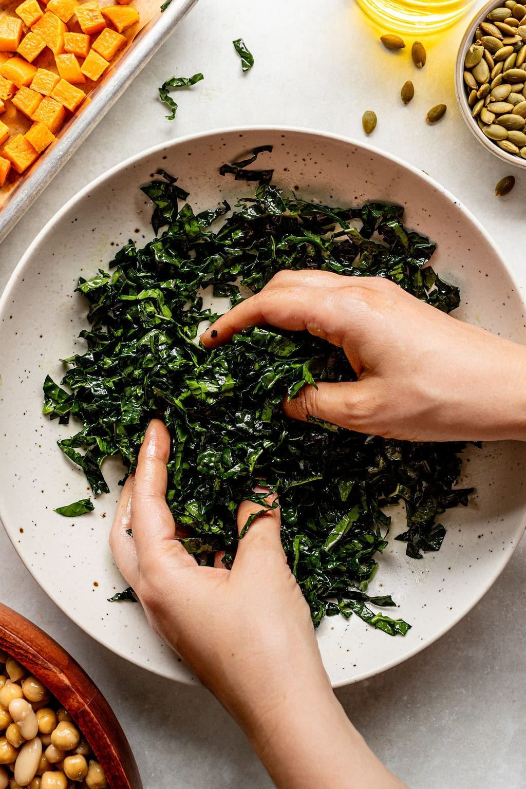 Two hands massage chopped kale in a white bowl, surrounded by bowls of chickpeas, pumpkin seeds, oil, and cubed sweet potatoes on a light surface.
