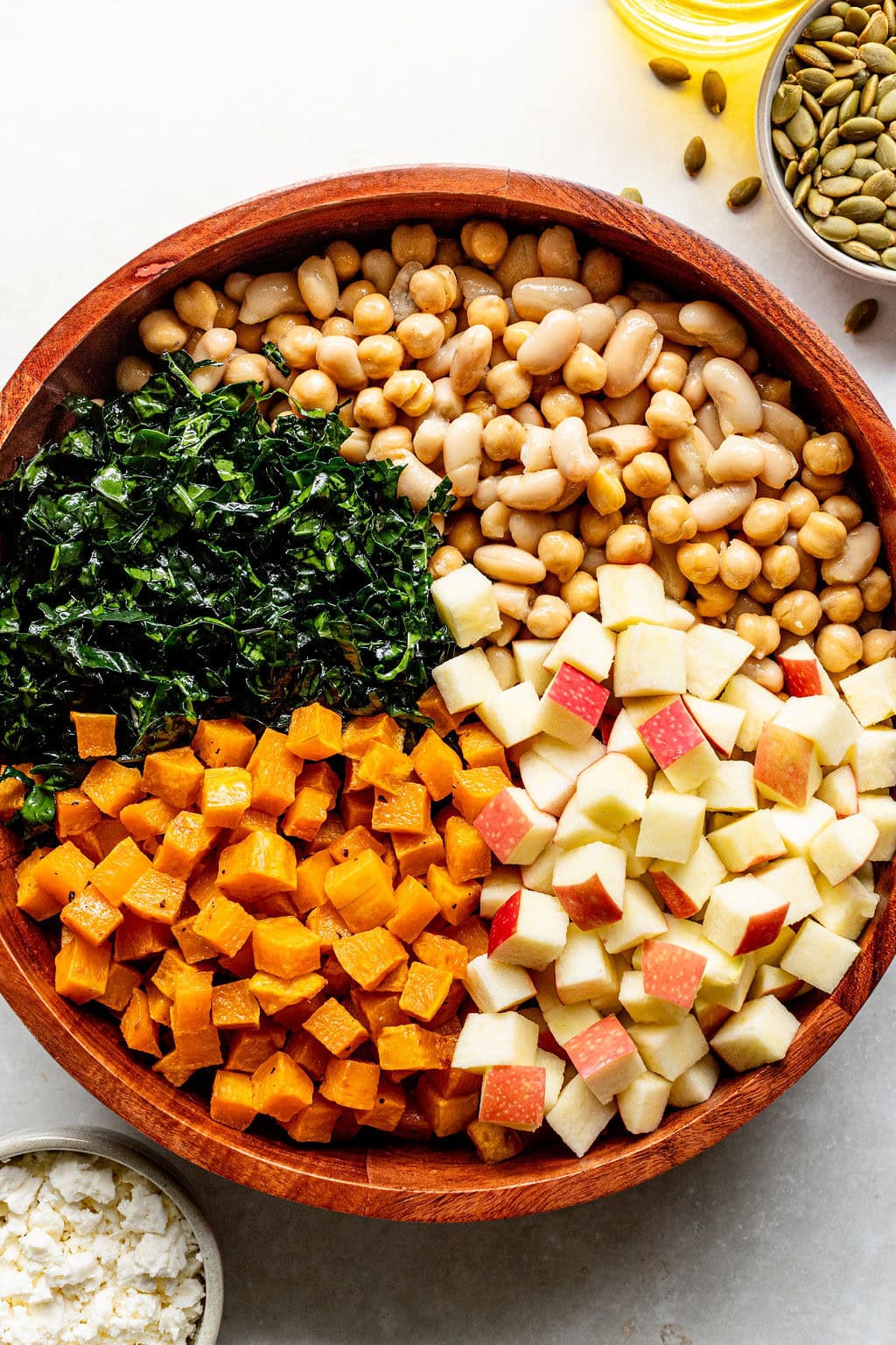 A wooden bowl filled with chopped kale, white beans, chickpeas, diced sweet potatoes, and diced red apples, shown from above.