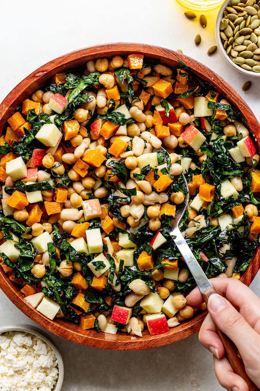 A hand holds a spoon in a wooden bowl filled with chopped kale, butternut squash, apples, beans, and chickpeas. Nearby are bowls of feta cheese, pumpkin seeds, and a bottle of oil.