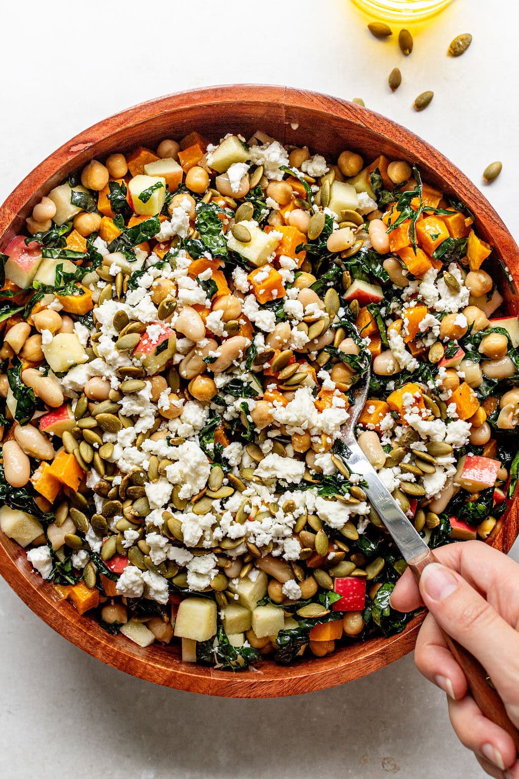 A hand holds a fork in a wooden bowl filled with a salad of beans, chopped vegetables, feta cheese, and seeds.