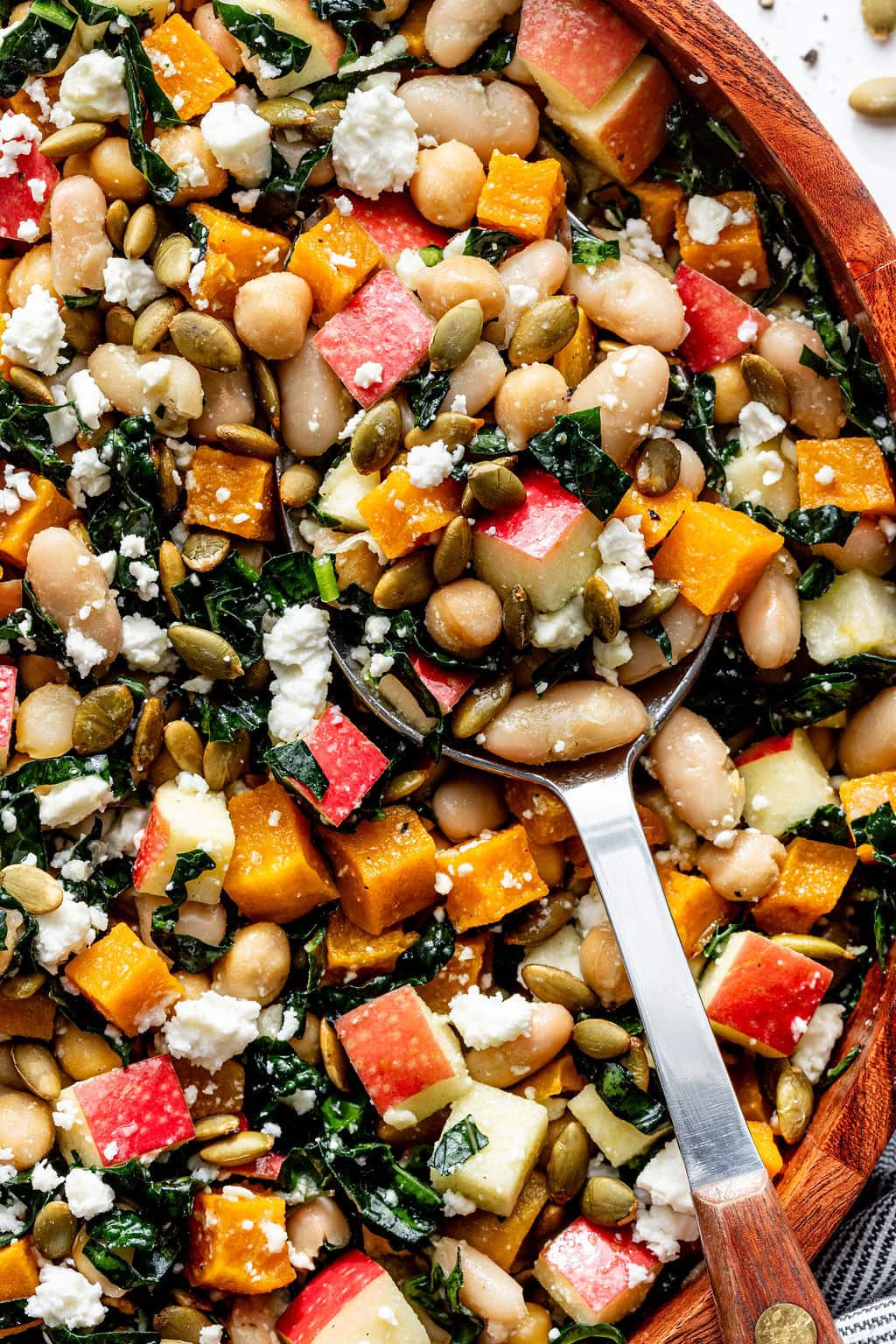 A close-up of a salad with white beans, diced apples, roasted butternut squash, kale, pumpkin seeds, and crumbled feta cheese, with a spoon resting in the bowl.