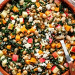A wooden bowl filled with salad containing kale, white beans, diced apples, roasted squash, pumpkin seeds, and crumbled cheese, with a serving spoon resting inside.