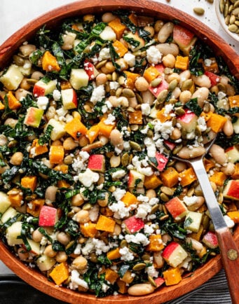 A wooden bowl filled with salad containing kale, white beans, diced apples, roasted squash, pumpkin seeds, and crumbled cheese, with a serving spoon resting inside.