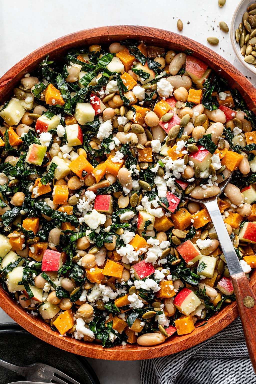 A wooden bowl filled with salad containing kale, white beans, diced apples, roasted squash, pumpkin seeds, and crumbled cheese, with a serving spoon resting inside.