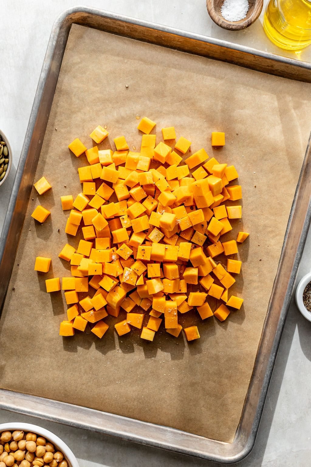 Diced butternut squash is spread on a parchment-lined baking sheet, surrounded by small bowls of oil, spices, seeds, and chickpeas.