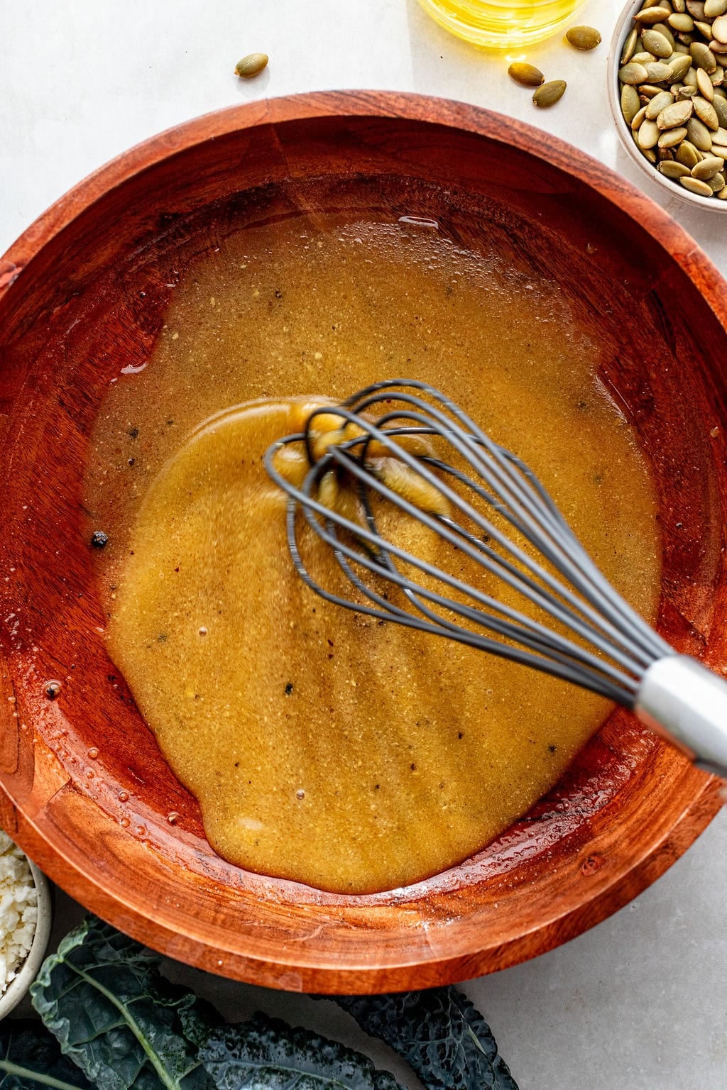 A metal whisk mixing a golden liquid dressing in a wooden bowl, with small bowls of seeds, oil, and leafy greens nearby.