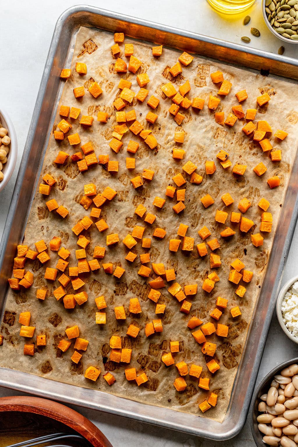 A baking sheet lined with parchment paper, topped with evenly spread roasted sweet potato cubes. Surrounding the sheet are small bowls filled with beans, seeds, and cheese.