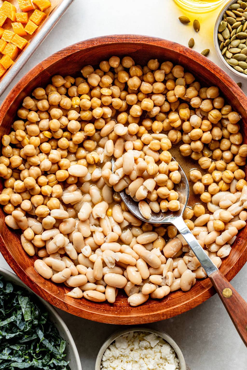 A wooden bowl filled with chickpeas and white beans, with a metal spoon, surrounded by small bowls of various salad ingredients.
