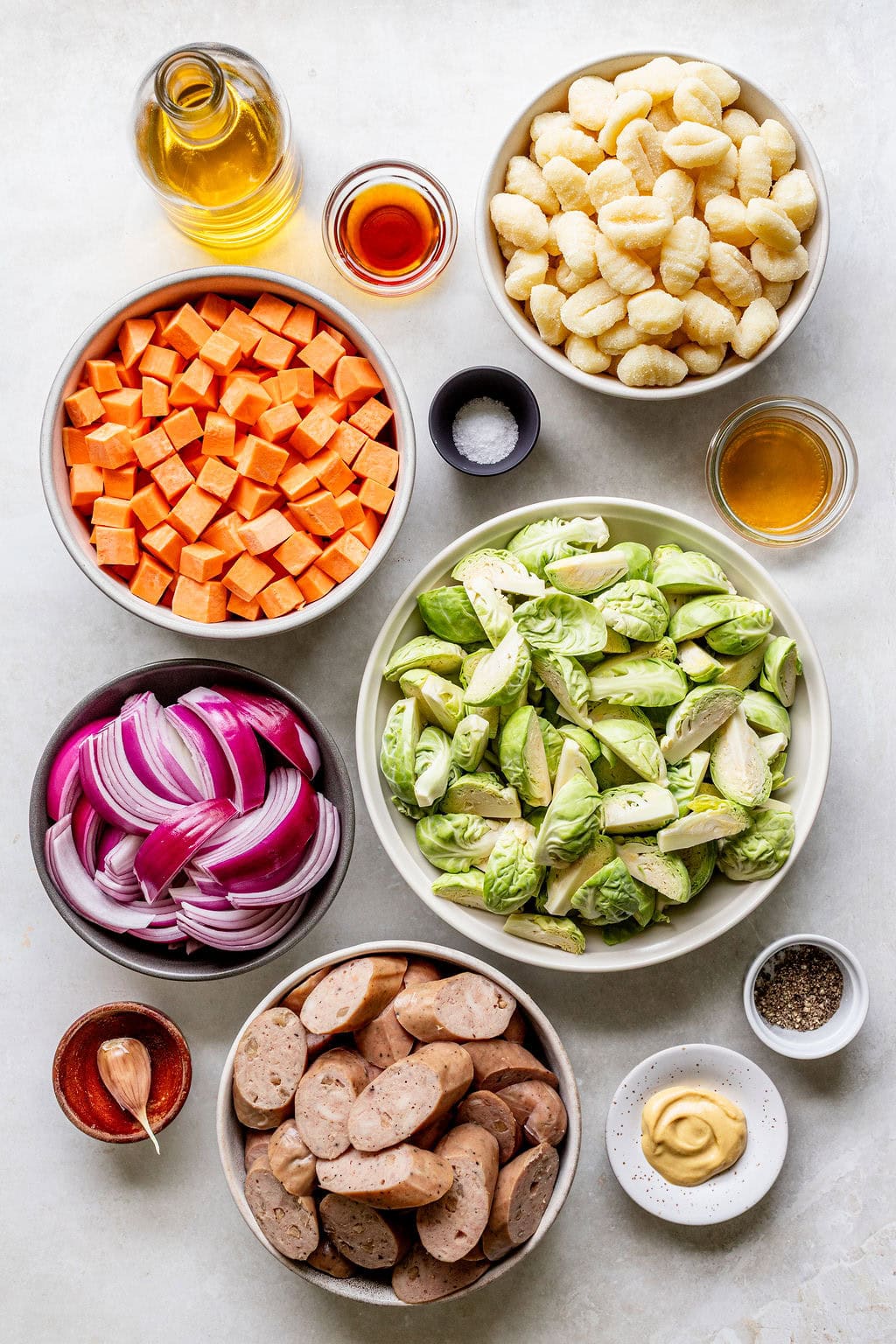 Bowls with gnocchi, diced sweet potatoes, halved Brussels sprouts, sliced sausages, red onion, garlic, olive oil, vinegar, mustard, salt, pepper, and honey arranged on a white surface.