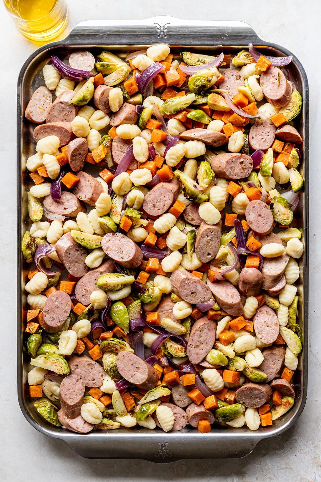 Sheet pan with sliced sausage, gnocchi, Brussels sprouts, red onion, and diced sweet potatoes, arranged and ready for roasting.