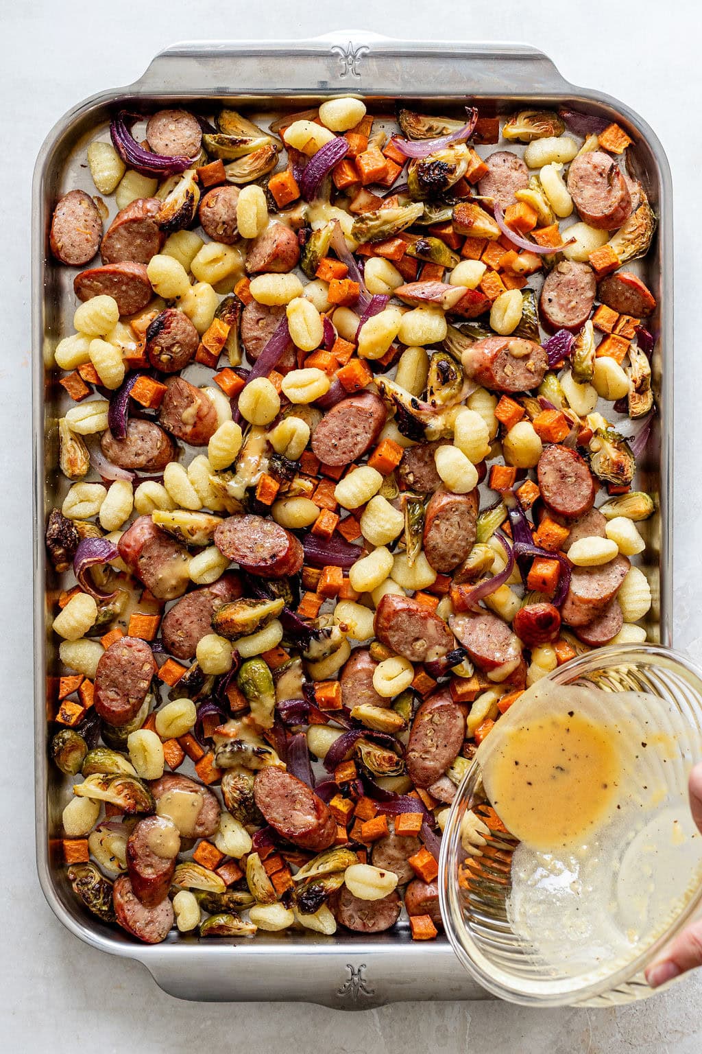 Sheet pan with cooked gnocchi, sausage, carrots, Brussels sprouts, and red onions; a hand pours sauce from a glass bowl over the mixture.