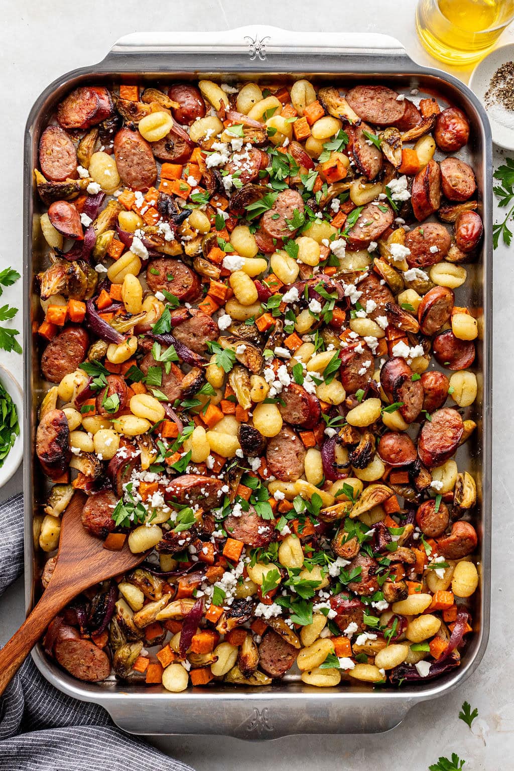 A sheet pan filled with roasted gnocchi, sausage slices, vegetables, and herbs, topped with crumbled cheese and fresh parsley, with a wooden spoon on the side.
