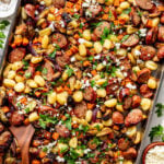 Sheet pan meal with sliced sausage, gnocchi, roasted vegetables, crumbled cheese, and fresh parsley, served in a large metal pan with a wooden spoon.