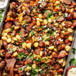 Sheet pan meal with sliced sausage, gnocchi, roasted vegetables, crumbled cheese, and fresh parsley, served in a large metal pan with a wooden spoon.