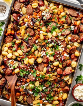 Sheet pan meal with sliced sausage, gnocchi, roasted vegetables, crumbled cheese, and fresh parsley, served in a large metal pan with a wooden spoon.