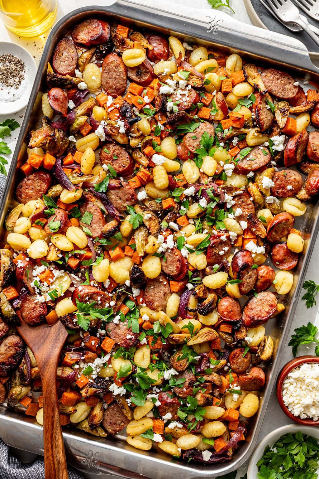 Sheet pan meal with sliced sausage, gnocchi, roasted vegetables, crumbled cheese, and fresh parsley, served in a large metal pan with a wooden spoon.