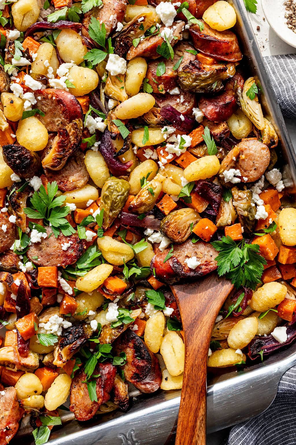 Sheet pan of roasted gnocchi, sausage, Brussels sprouts, sweet potatoes, and red onions, garnished with chopped parsley and crumbled cheese, with a wooden spoon in the tray.