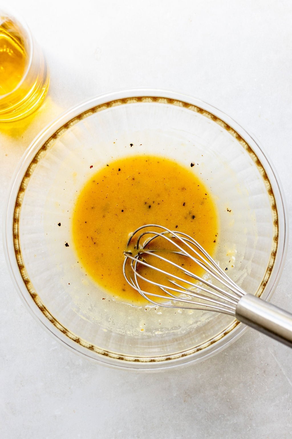 A metal whisk in a glass bowl mixes a yellow vinaigrette dressing with visible black pepper. A small glass of oil sits nearby on a white surface.