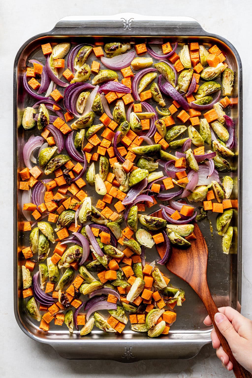 A baking sheet filled with roasted Brussels sprouts, cubed sweet potatoes, and red onion slices; a hand holds a wooden spoon, stirring the vegetables.