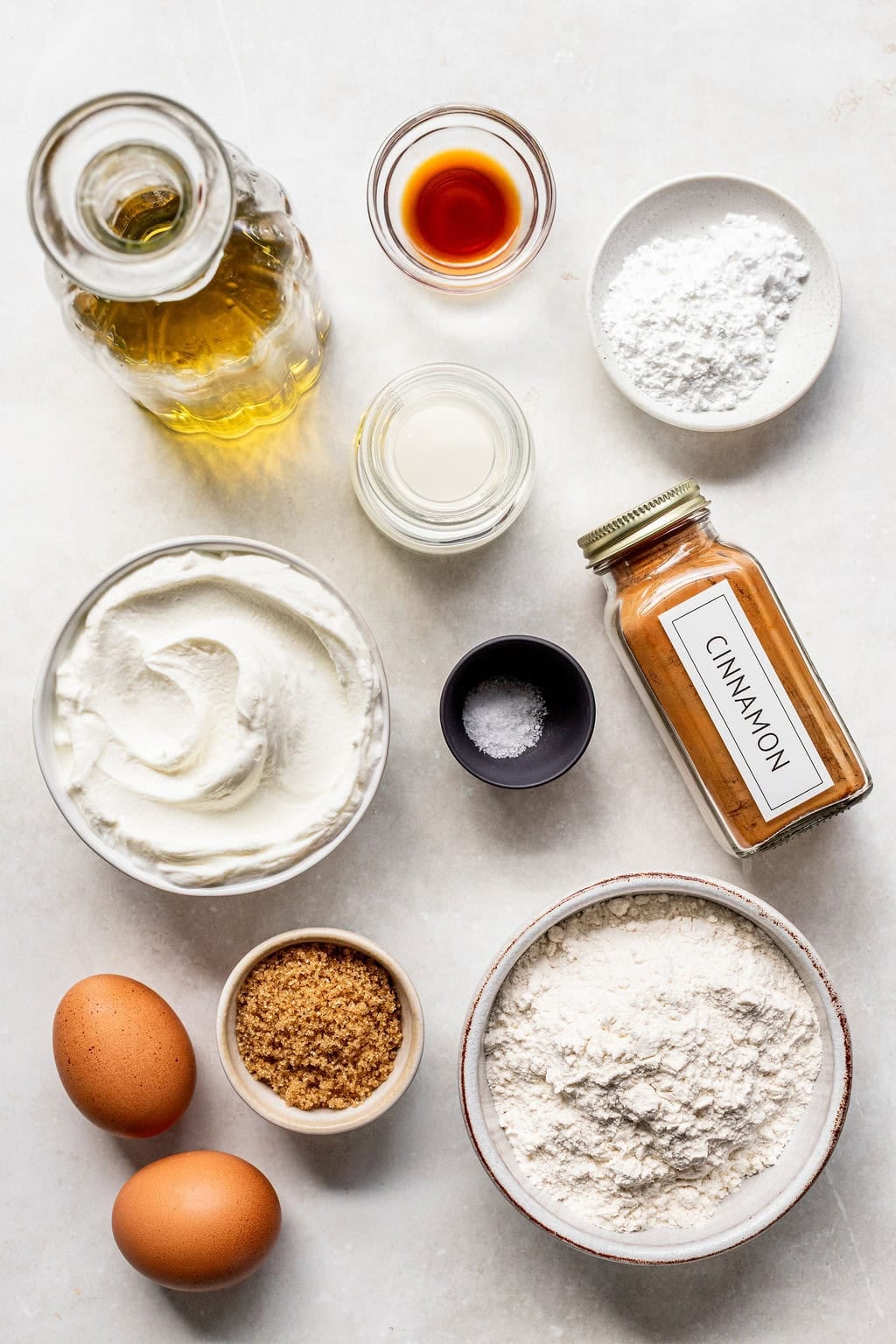 Various baking ingredients, including eggs, brown sugar, flour, yogurt, cinnamon, vanilla, oil, salt, and powdered sugar, arranged in bowls and jars on a white surface.