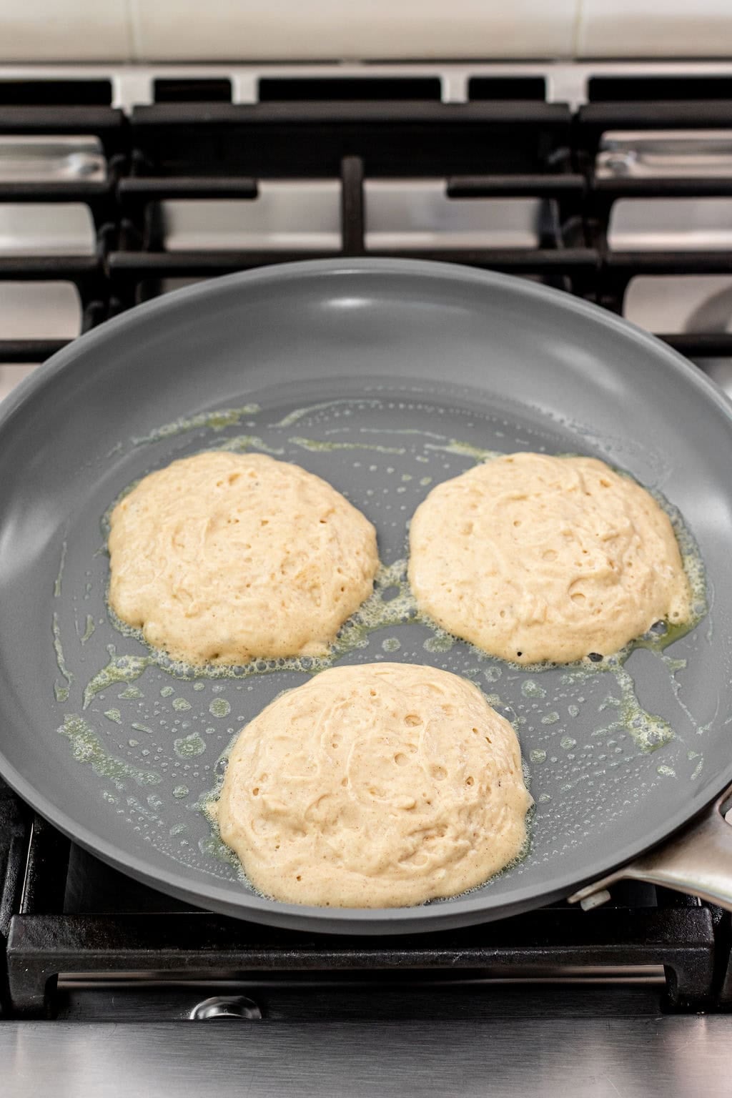 Three pancakes are cooking in a greased nonstick skillet on a stovetop, with bubbles forming on their surfaces.