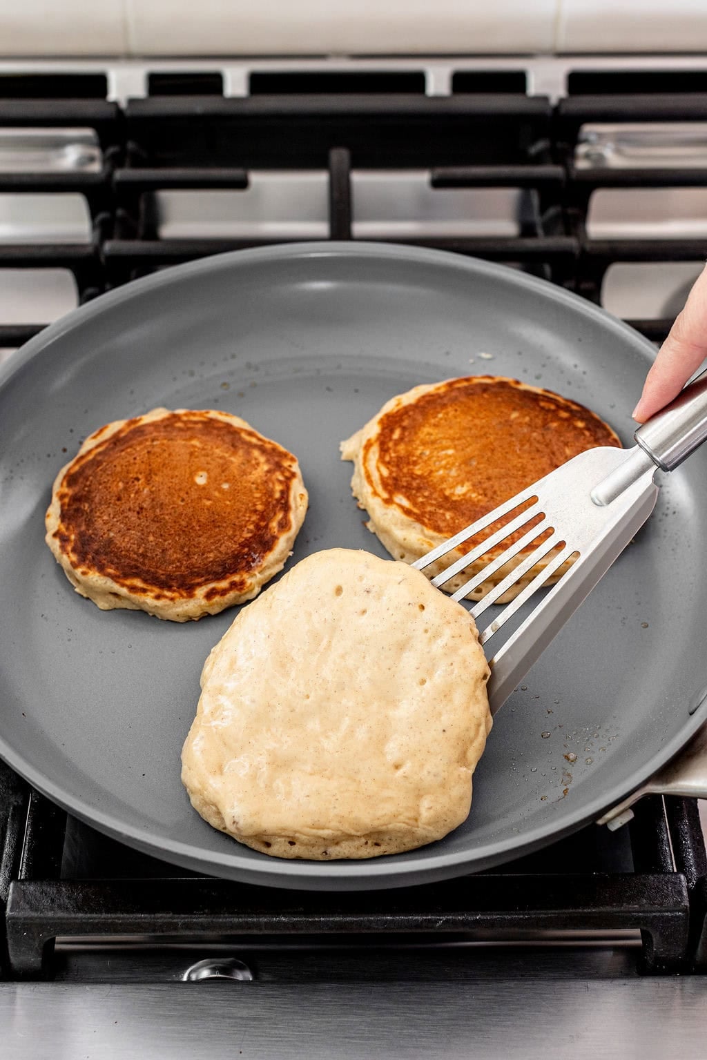 A spatula lifts a pancake in a frying pan, while two cooked pancakes sit nearby in the pan.