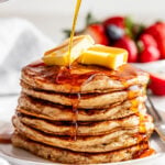 A stack of pancakes topped with butter is being drizzled with syrup, with strawberries and blueberries in the background.