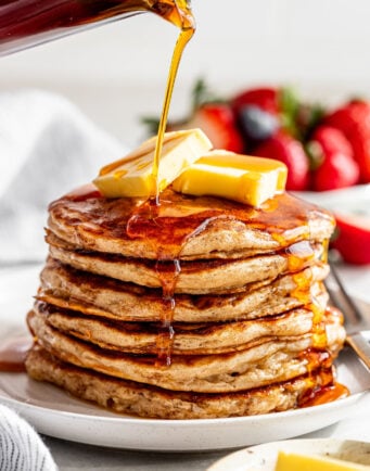 A stack of pancakes topped with butter is being drizzled with syrup, with strawberries and blueberries in the background.