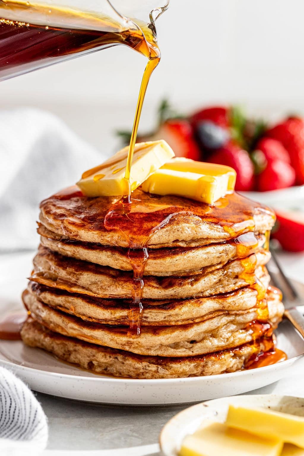 A stack of pancakes topped with butter is being drizzled with syrup, with strawberries and blueberries in the background.